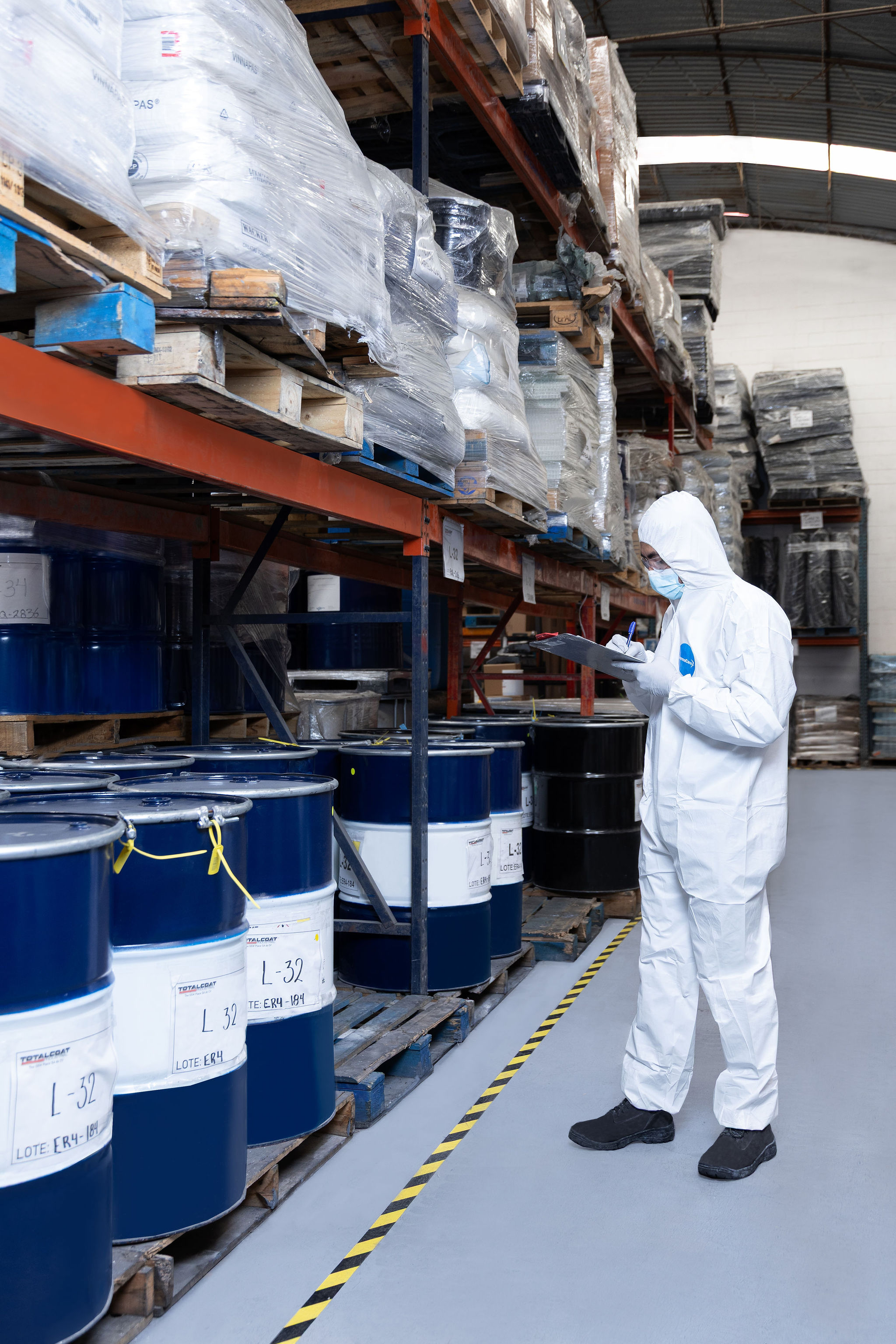 Técnico con traje de protección revisando inventario de tambores en almacén.